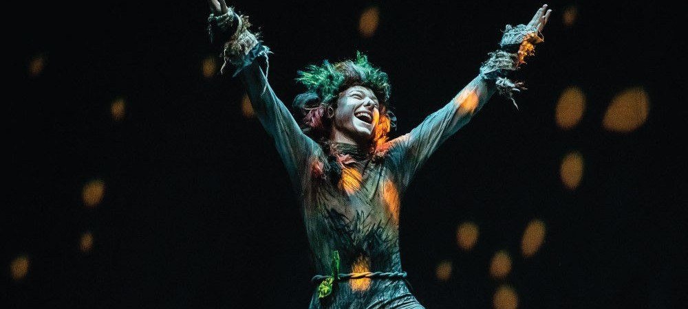 A performer in a green costume, arms raised, on a dark stage